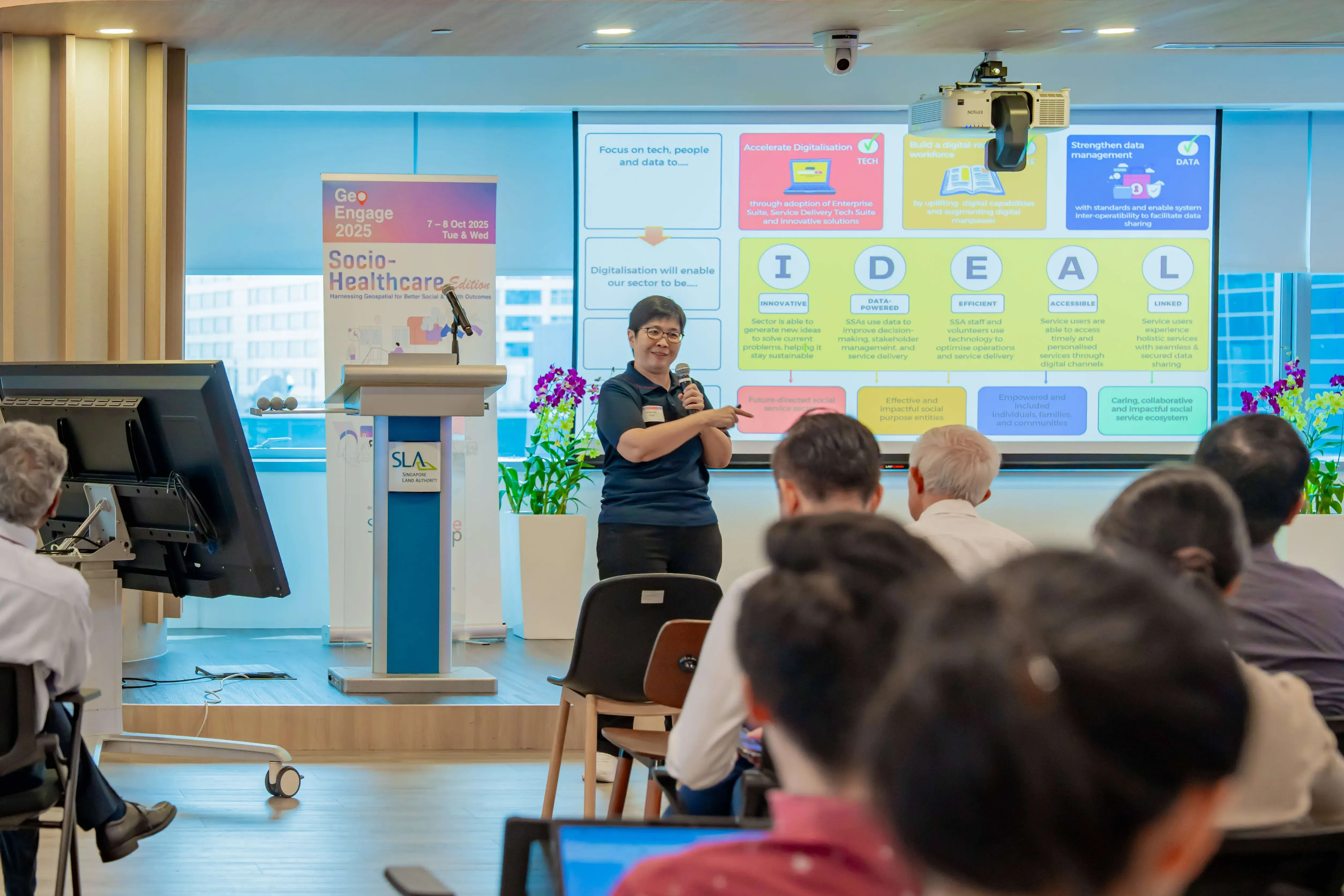 A speaker holds a microphone at a GeoEngage 2025 Socio-Healthcare conference, presenting an IDEAL framework slide to seated attendees.